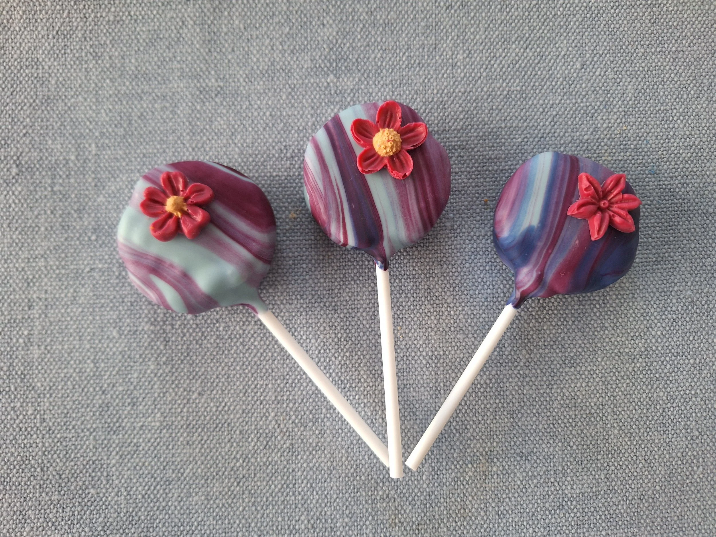 Three marbled cake pops in pink and purple tones, each decorated with a small flower