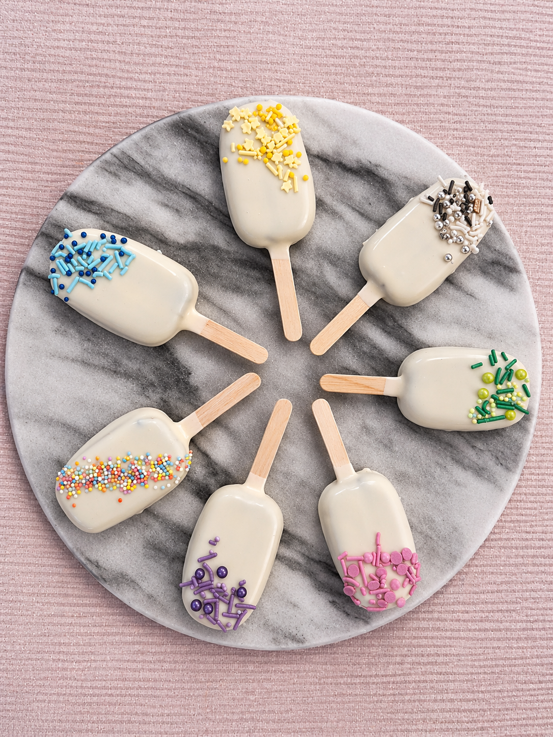 Seven white cakesicles with colourful sprinkles arranged in a circle on a marble-patterned plate