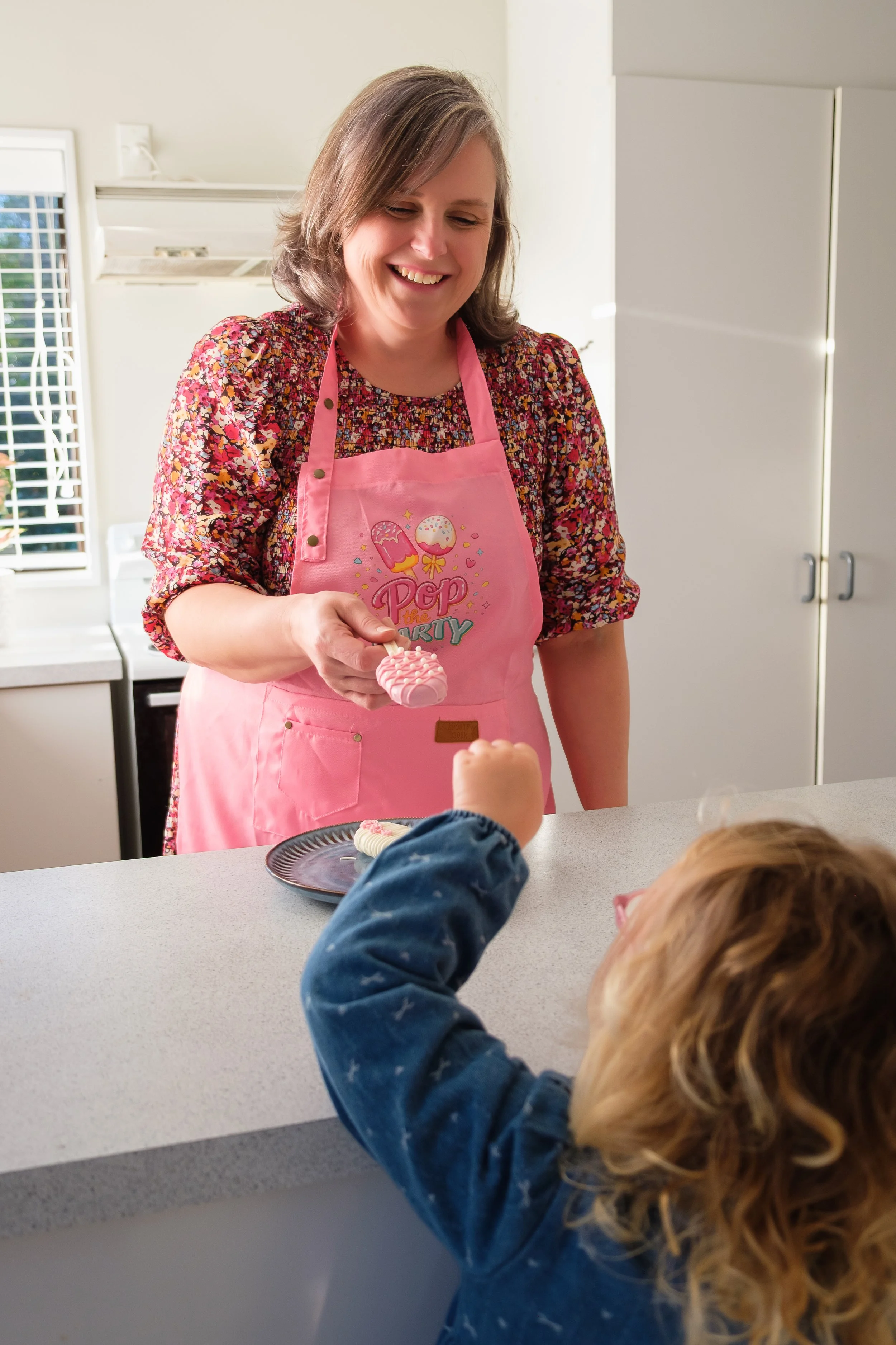 Eve in a pink apron sharing a treat with a child across the kitchen counter